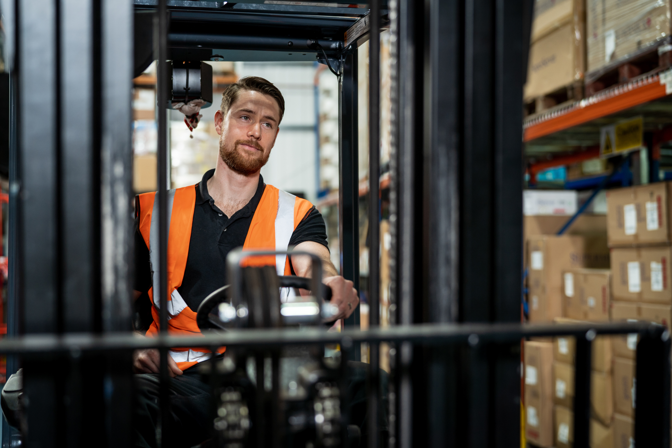 Man wearing an orange vest in a manufacturing or warehouse driving a forklift. Key words: tele conferencing, cloud calling, Microsoft Teams, meeting, conference call, phone system, video system, Business Voice, Manufacturing, Firstline Worker, Delivery, Tracking