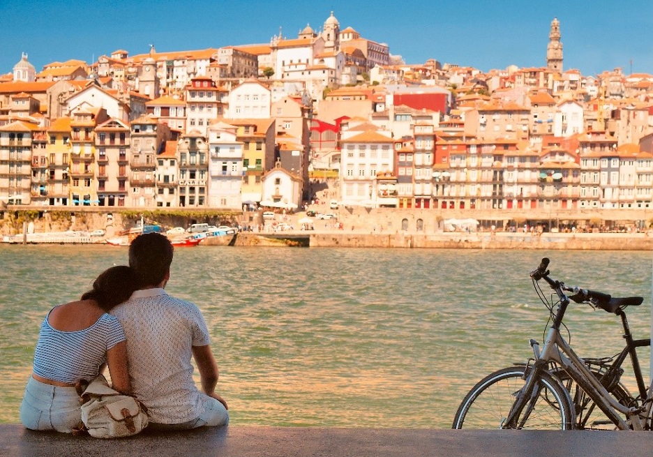 A couple sit together on the bank of a river, looking across at a Portuguese town on the opposite bank